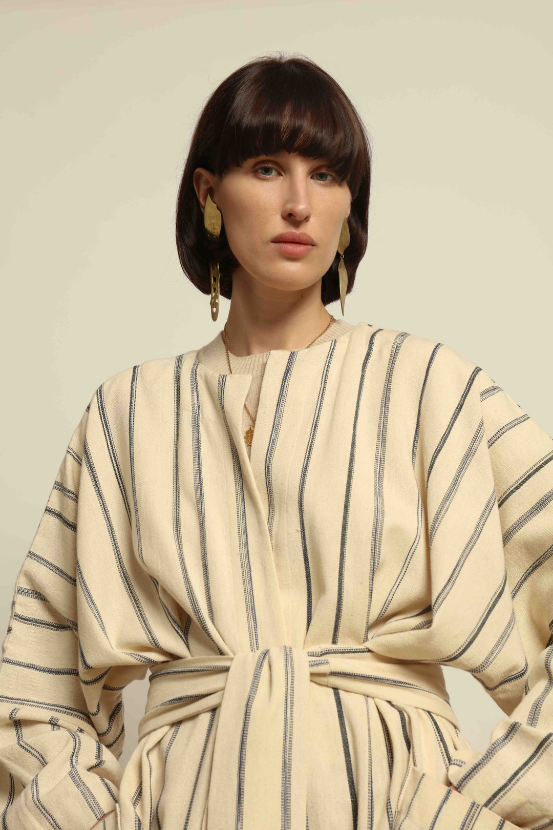Model is wearing the Maxi Handwoven and Hand-Printed Benin Cotton Kimono ‘The Ones Who Laugh’ by Atelier V.O, crafted from 100% handwoven cotton with Dogon Mali-inspired print, straight cut, drawstring, wooden button fastening, and matching belt.