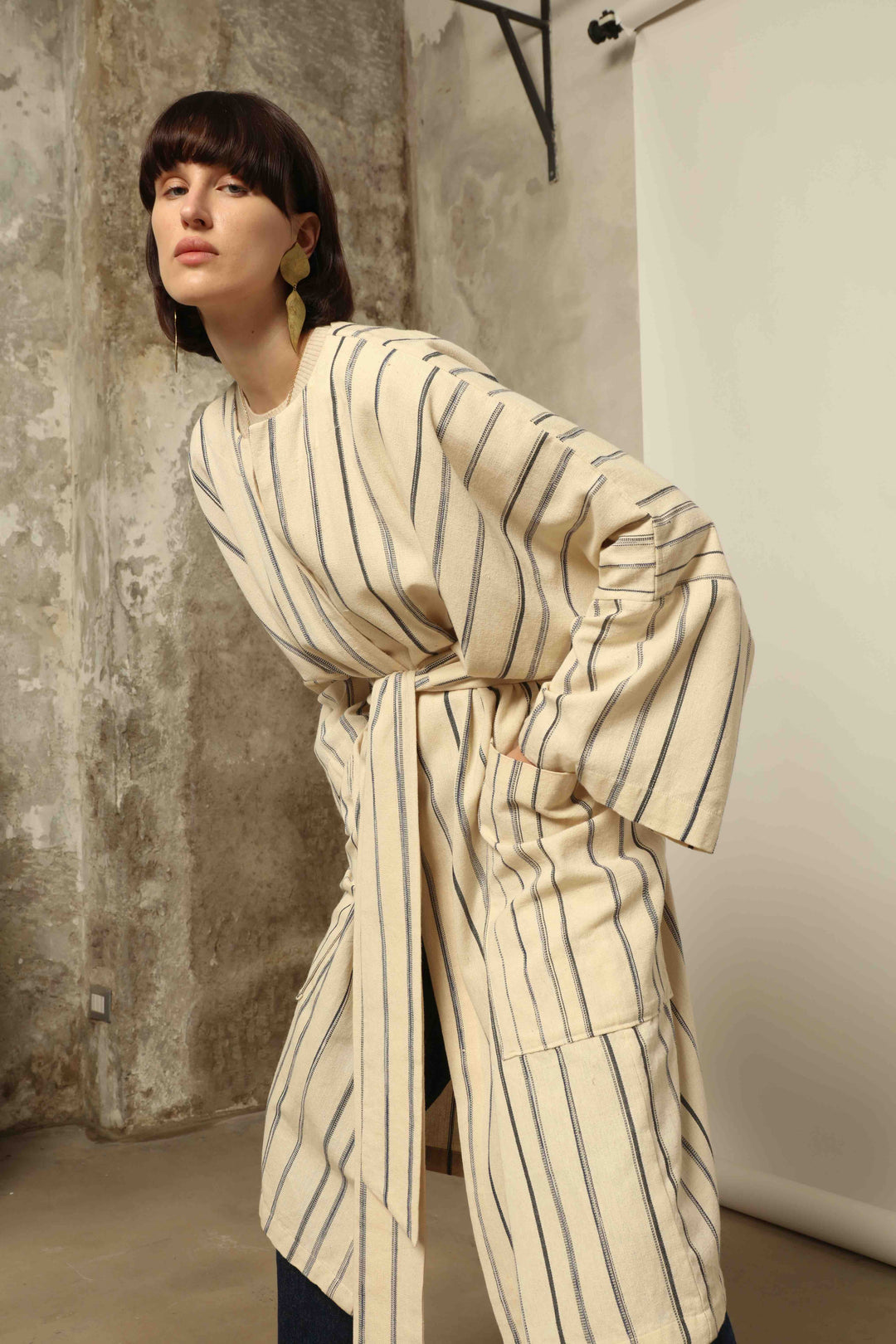 Model is wearing the Maxi Handwoven and Hand-Printed Benin Cotton Kimono ‘The Ones Who Laugh’ by Atelier V.O, crafted from 100% handwoven cotton with Dogon Mali-inspired print, straight cut, drawstring, wooden button fastening, and matching belt.