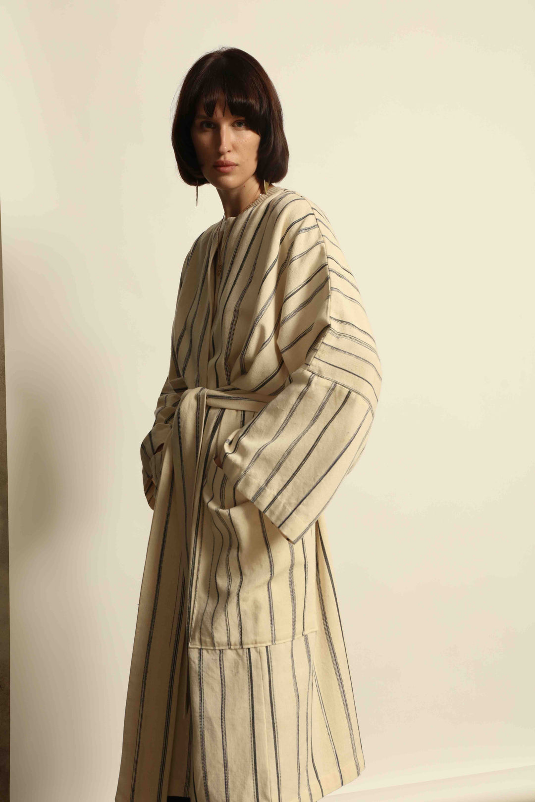 Model is wearing the Maxi Handwoven and Hand-Printed Benin Cotton Kimono ‘The Ones Who Laugh’ by Atelier V.O, crafted from 100% handwoven cotton with Dogon Mali-inspired print, straight cut, drawstring, wooden button fastening, and matching belt.