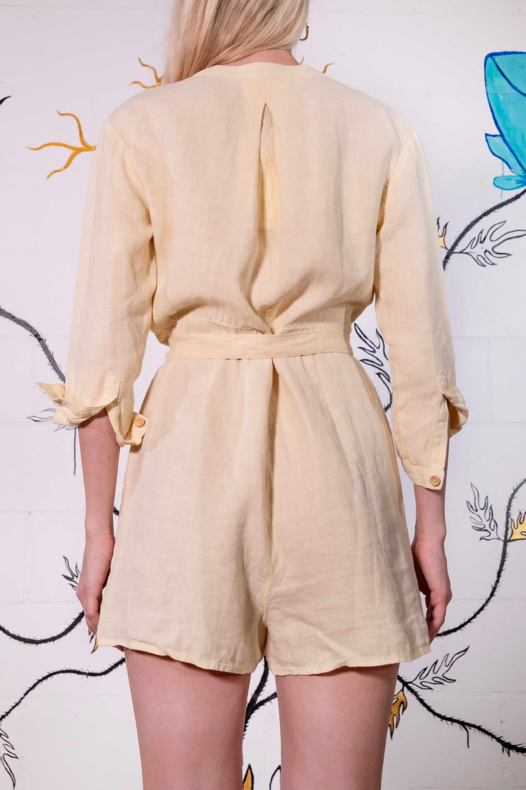 Model is wearing a banana yellow short linen jumpsuit by Happy Haus, featuring a relaxed fit, oversized chest and side pockets, wooden buttons, and a matching fabric belt.