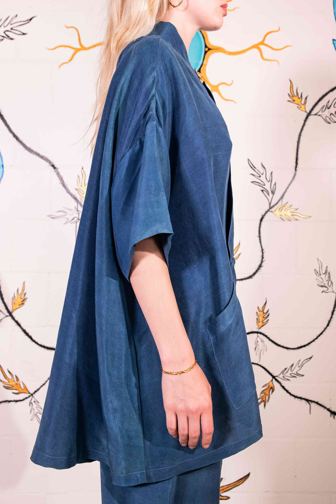 Model is wearing a short-sleeve indigo shirt made from 100% silk, featuring a relaxed fit, dropped shoulders, front button closure, and large patch pockets. Hand-dyed using botanical pigments by A Sacred Wrap.