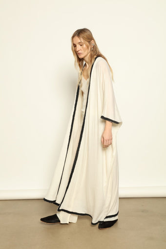 Natural White Long Cardigan by Mirror in the Sky crafted from 100% pure undyed cashmere. Woman wearing a long white dress with black trim on a plain background