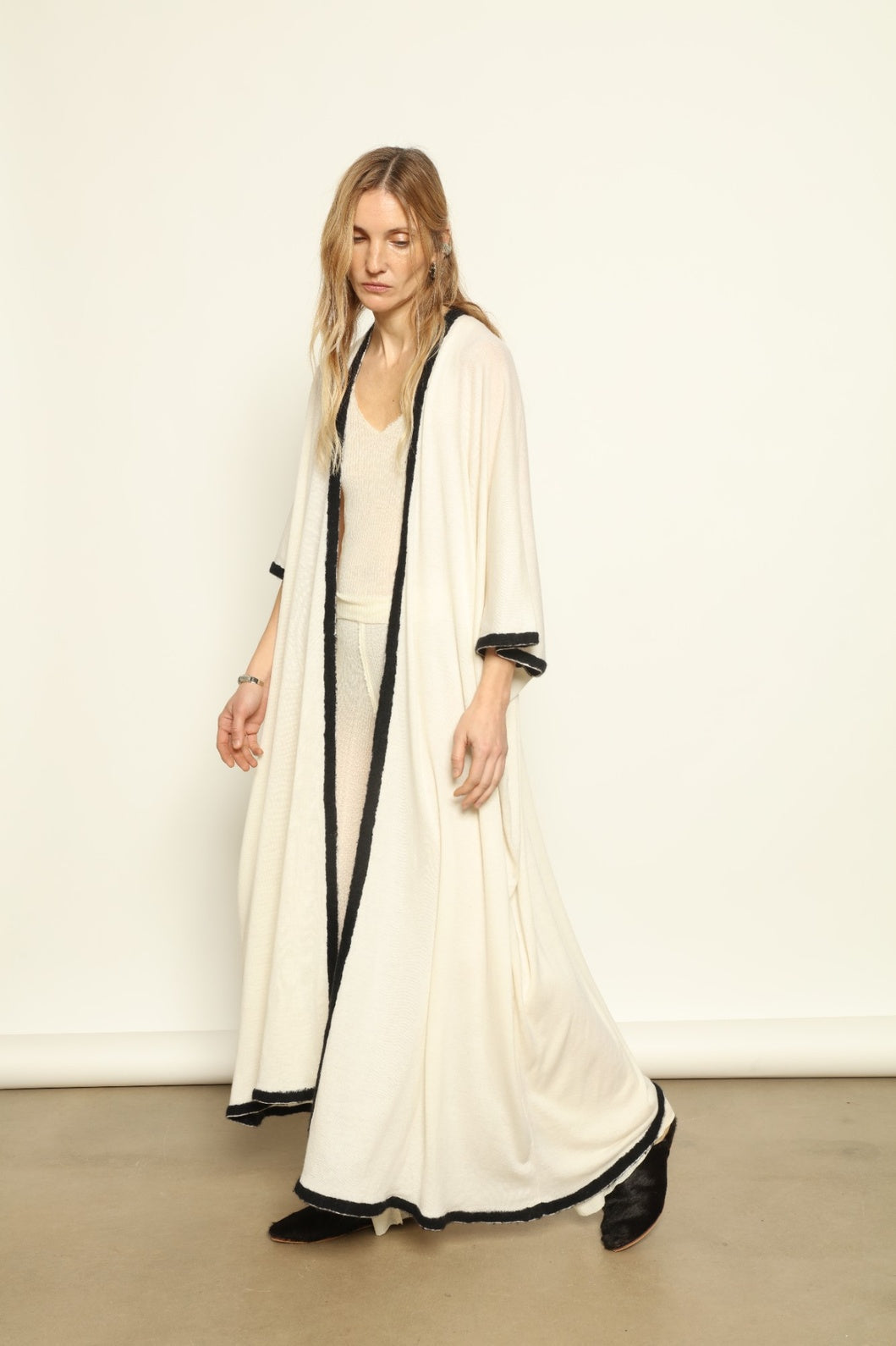 Natural white cashmere cardigan by Mirror in the Sky offering lightweight warmth and fluid movement. Woman wearing a long white dress with black trim against a plain background