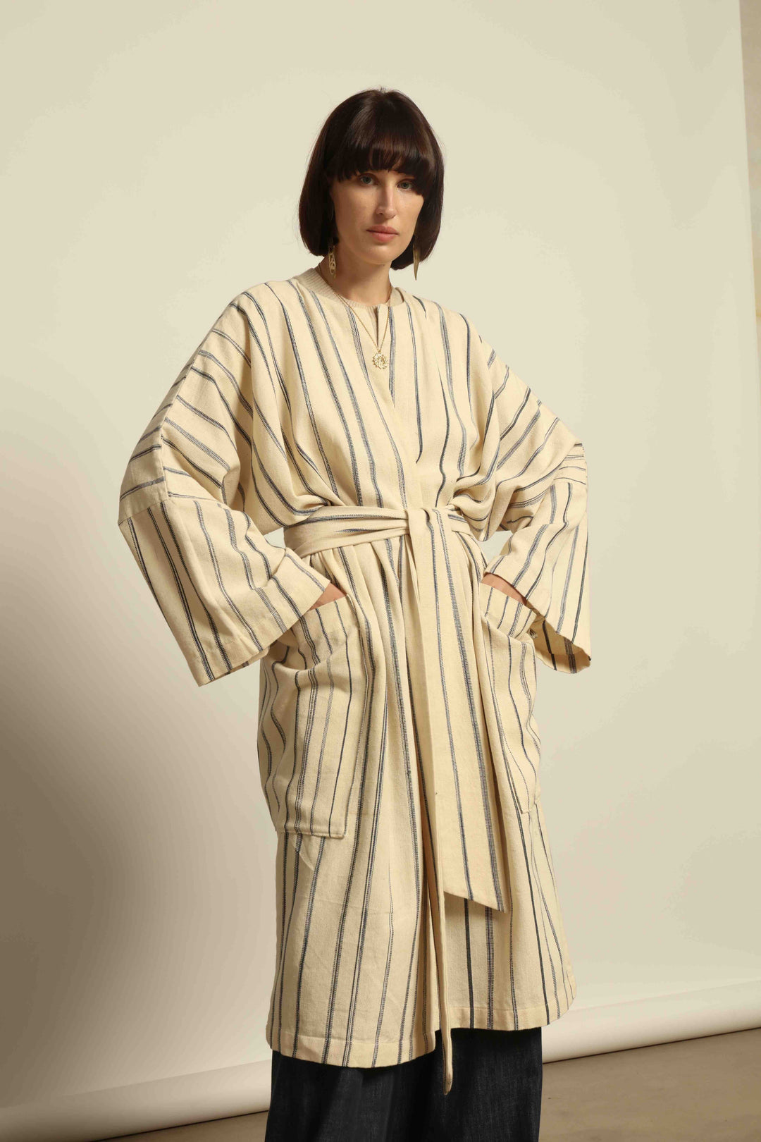 Model is wearing the Maxi Handwoven and Hand-Printed Benin Cotton Kimono ‘The Ones Who Laugh’ by Atelier V.O, crafted from 100% handwoven cotton with Dogon Mali-inspired print, straight cut, drawstring, wooden button fastening, and matching belt.