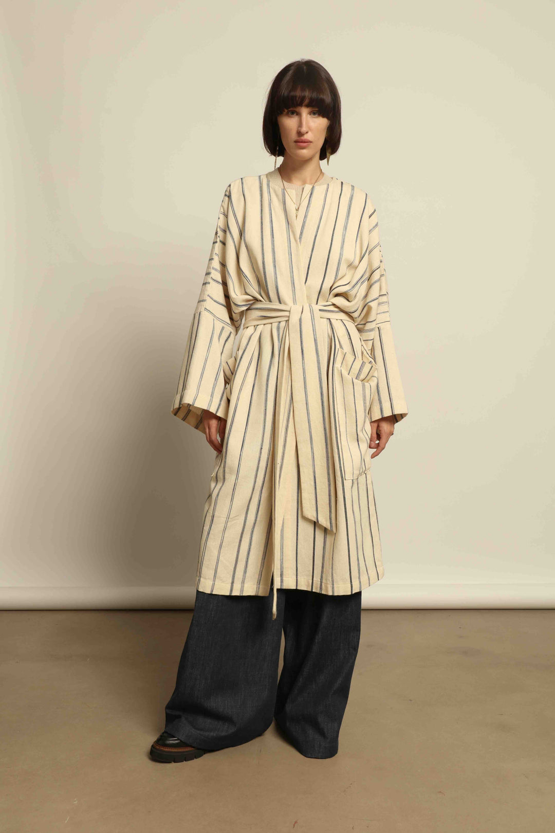 Model is wearing the Maxi Handwoven and Hand-Printed Benin Cotton Kimono ‘The Ones Who Laugh’ by Atelier V.O, crafted from 100% handwoven cotton with Dogon Mali-inspired print, straight cut, drawstring, wooden button fastening, and matching belt.