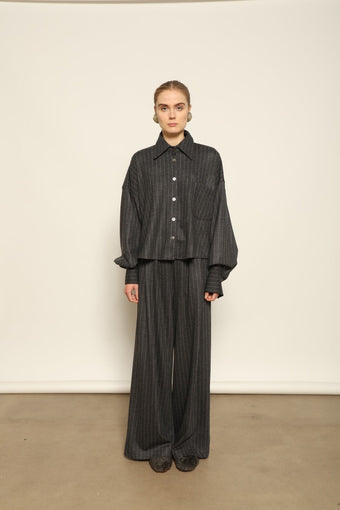 Model is wearing Bettina Striped Grey and White Double Jersey Shirt by House of Nomad Stories, part of the curated selection, handcrafted in Italy from linen, wool, and silk blend with vertical stripes, relaxed tailoring, and fluid drape for timeless elegance.