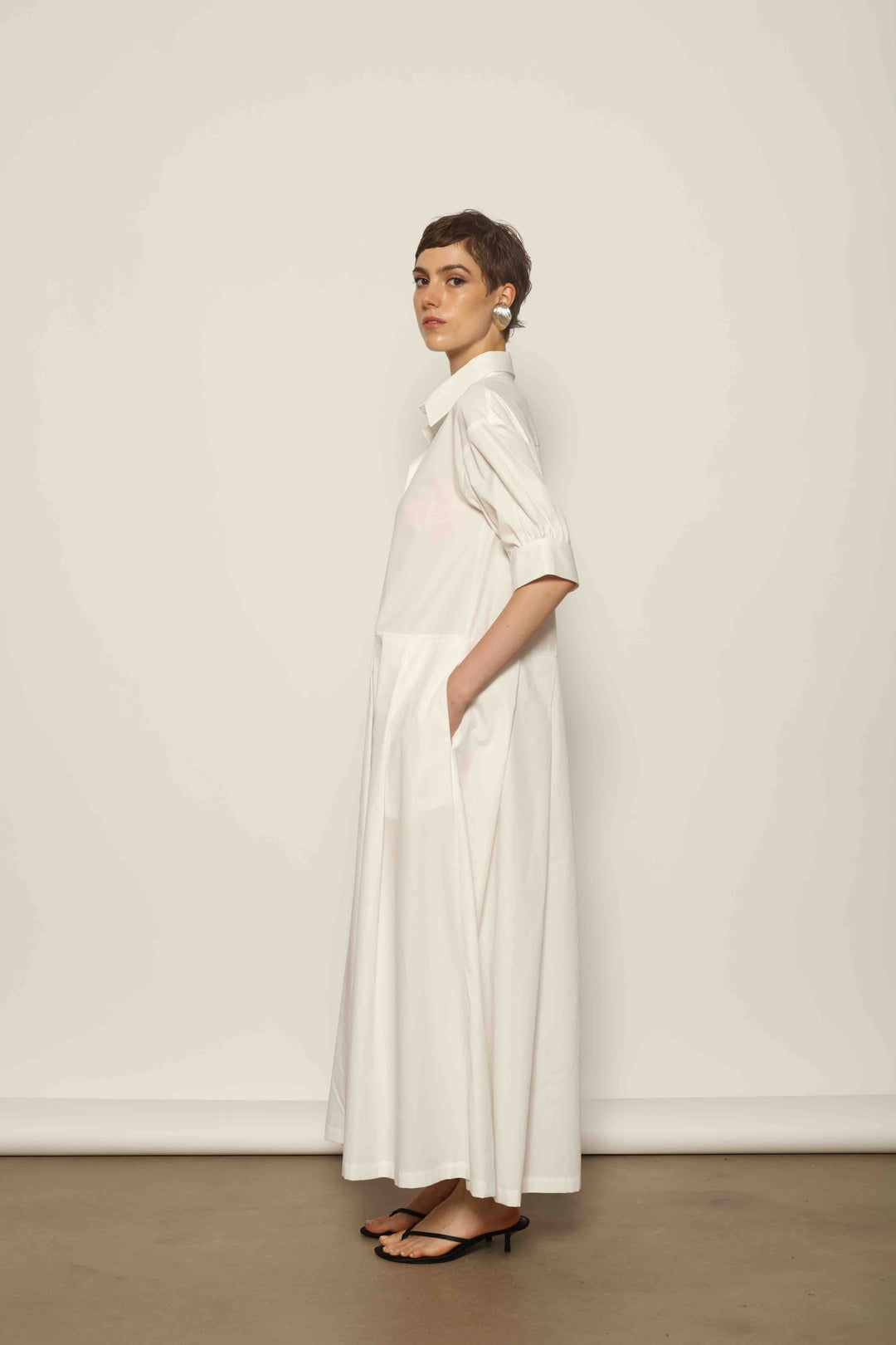 Model wearing the Emily Organic White Cotton Dress by House of Nomad Stories, featuring 100% organic cotton fabric, classic collar, button-down front, and adjustable belt for a clean, elegant silhouette.