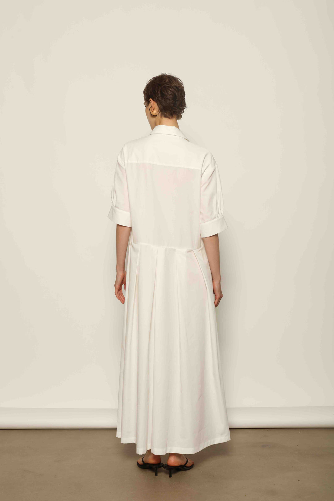 Model wearing the Emily Organic White Cotton Dress by House of Nomad Stories, featuring 100% organic cotton fabric, classic collar, button-down front, and adjustable belt for a clean, elegant silhouette.