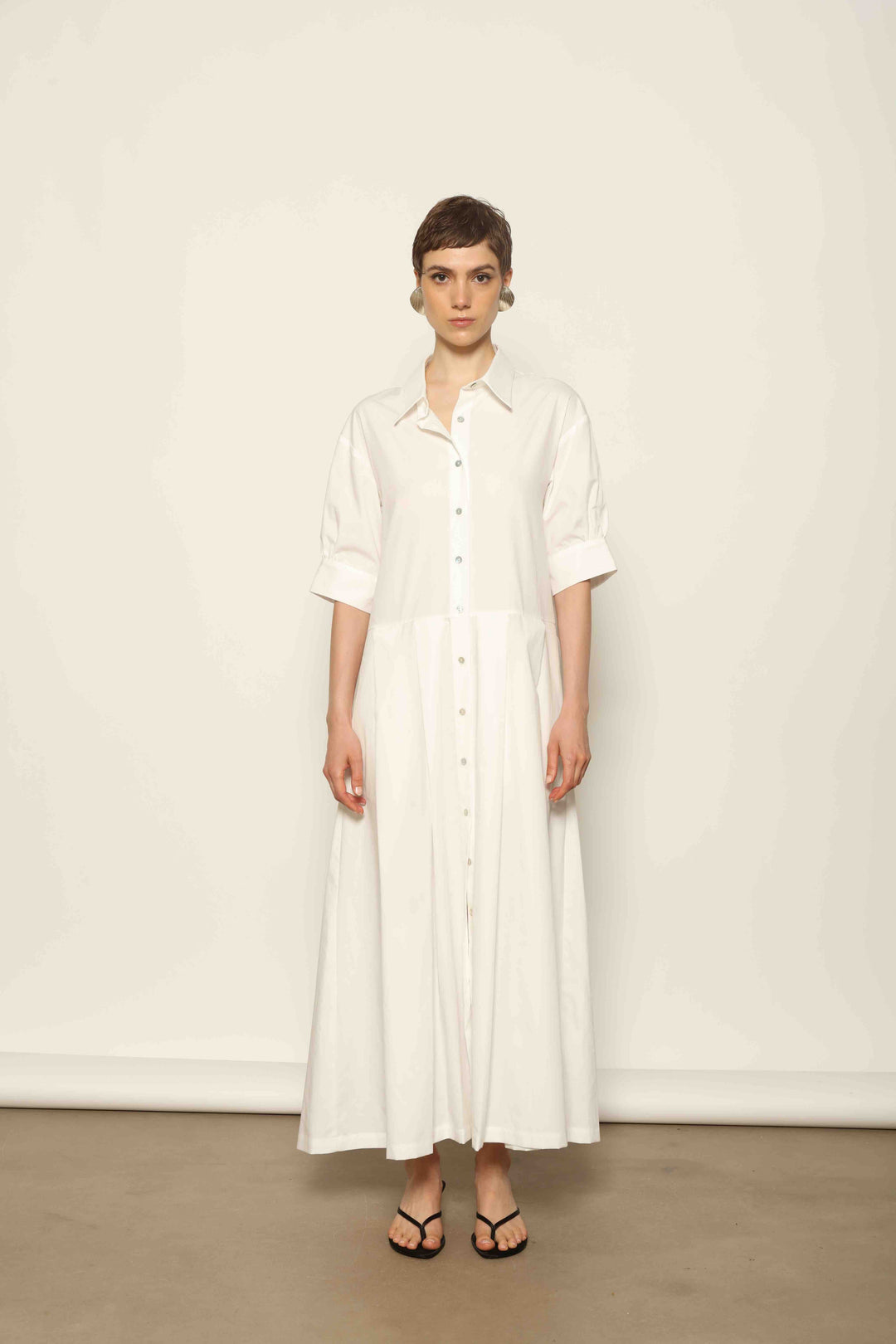 Model wearing the Emily Organic White Cotton Dress by House of Nomad Stories, featuring 100% organic cotton fabric, classic collar, button-down front, and adjustable belt for a clean, elegant silhouette.