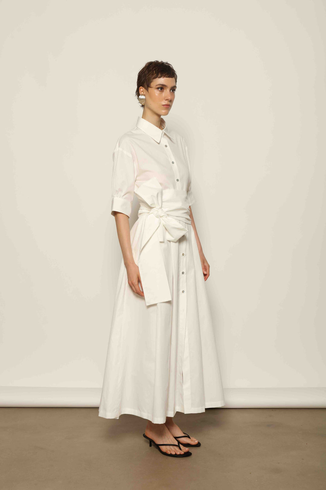 Model wearing the Emily Organic White Cotton Dress by House of Nomad Stories, featuring 100% organic cotton fabric, classic collar, button-down front, and adjustable belt for a clean, elegant silhouette.