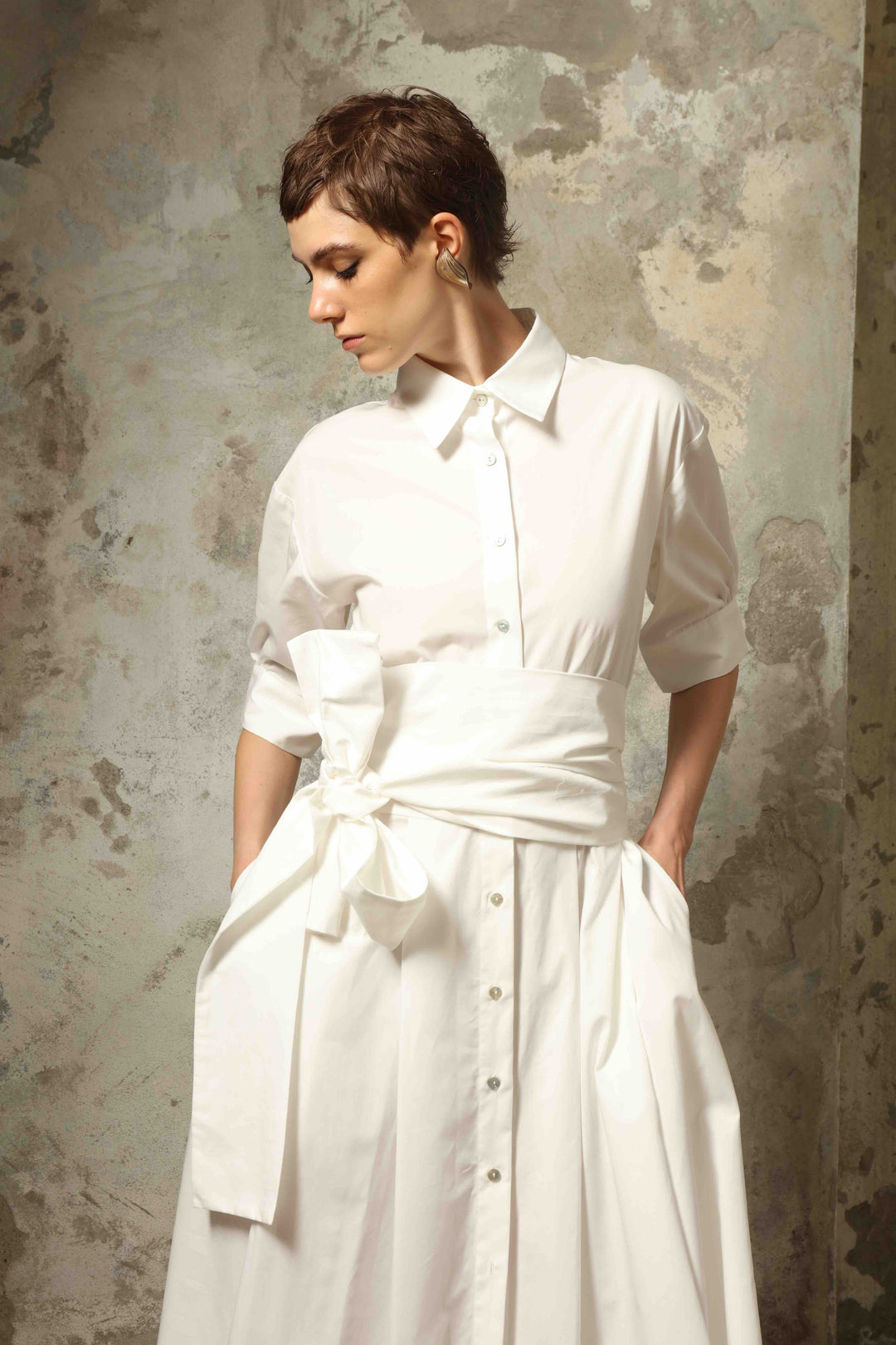 Model wearing the Emily Organic White Cotton Dress by House of Nomad Stories, featuring 100% organic cotton fabric, classic collar, button-down front, and adjustable belt for a clean, elegant silhouette.