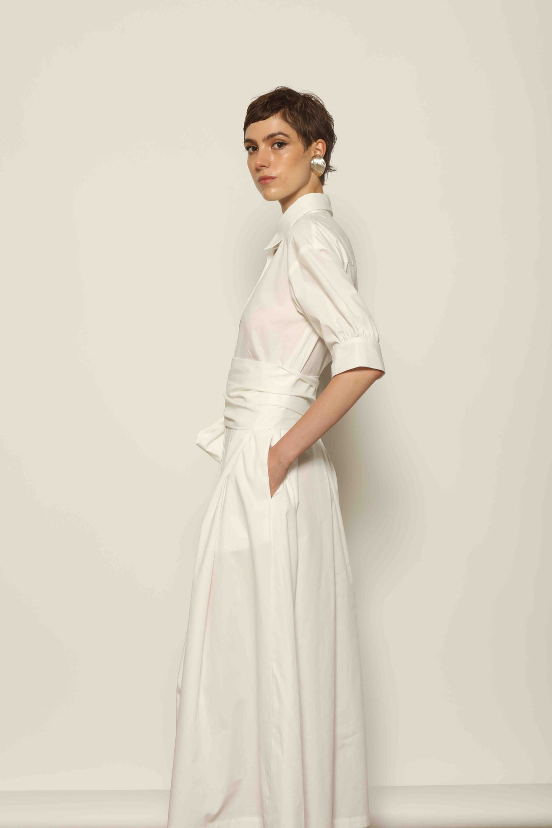 Model wearing the Emily Organic White Cotton Dress by House of Nomad Stories, featuring 100% organic cotton fabric, classic collar, button-down front, and adjustable belt for a clean, elegant silhouette.