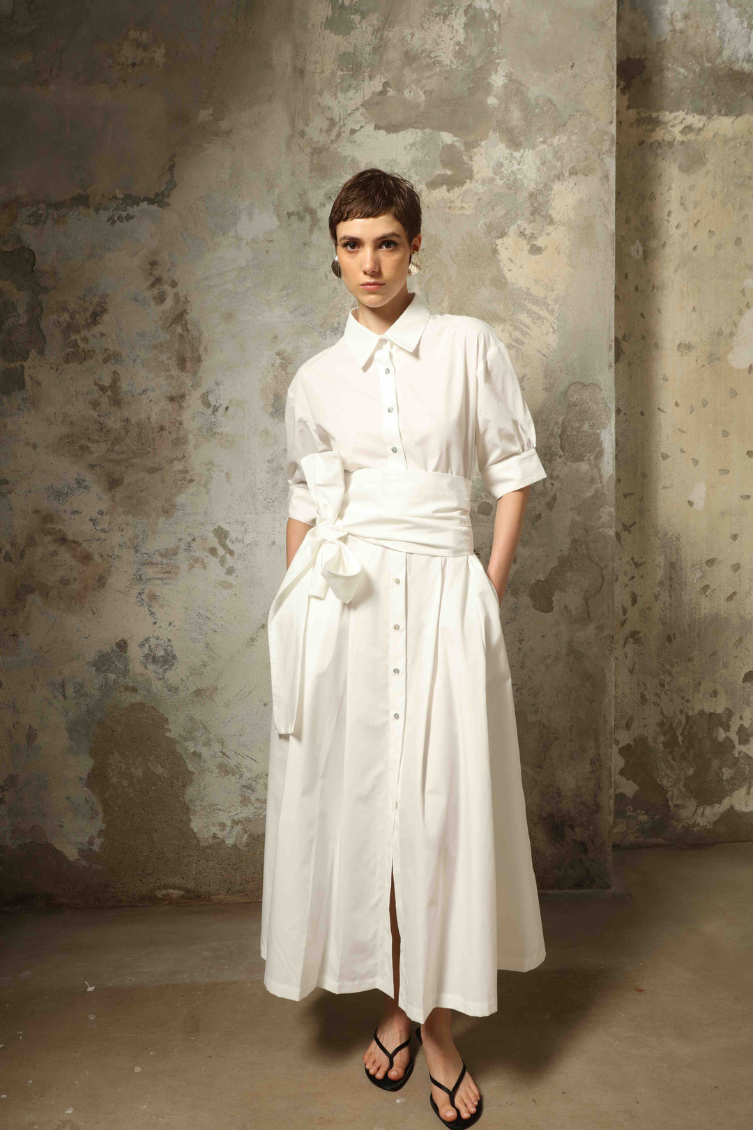 Model wearing the Emily Organic White Cotton Dress by House of Nomad Stories, featuring 100% organic cotton fabric, classic collar, button-down front, and adjustable belt for a clean, elegant silhouette.