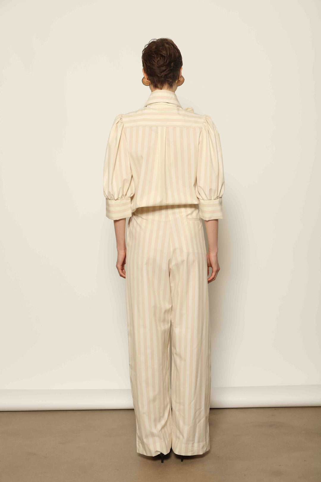 Model wearing the Gabriella Sand White Striped Trousers by House of Nomad Stories, featuring a high-rise waist, straight-leg tailored fit, and soft sand and white stripe pattern in a premium cotton, merino wool, and cashmere blend.