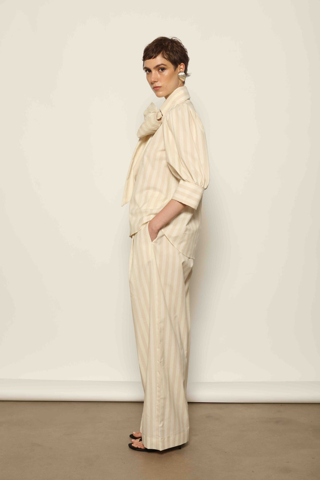 Model wearing the Gabriella Sand White Striped Trousers by House of Nomad Stories, featuring a high-rise waist, straight-leg tailored fit, and soft sand and white stripe pattern in a premium cotton, merino wool, and cashmere blend.