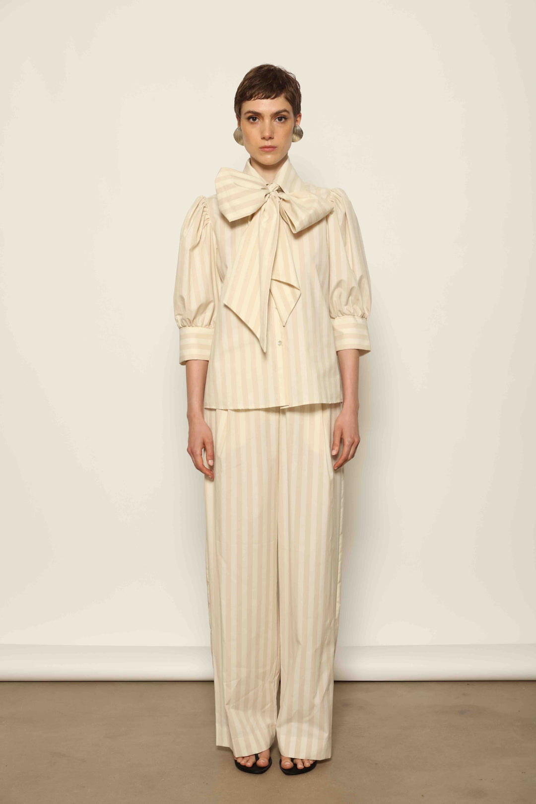 Model wearing the Gabriella Sand White Striped Trousers by House of Nomad Stories, featuring a high-rise waist, straight-leg tailored fit, and soft sand and white stripe pattern in a premium cotton, merino wool, and cashmere blend.