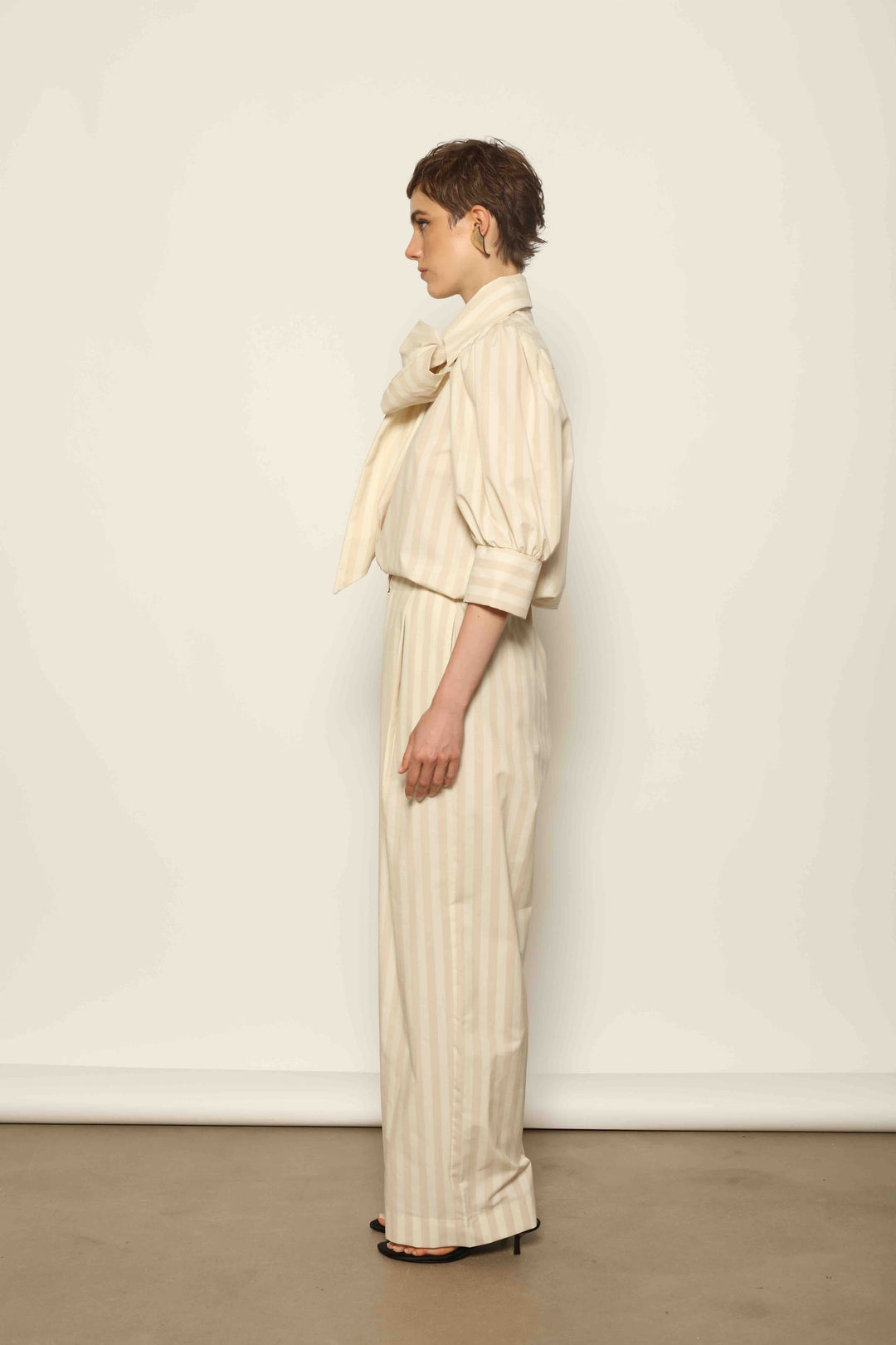 Model wearing the Gabriella Sand White Striped Trousers by House of Nomad Stories, featuring a high-rise waist, straight-leg tailored fit, and soft sand and white stripe pattern in a premium cotton, merino wool, and cashmere blend.