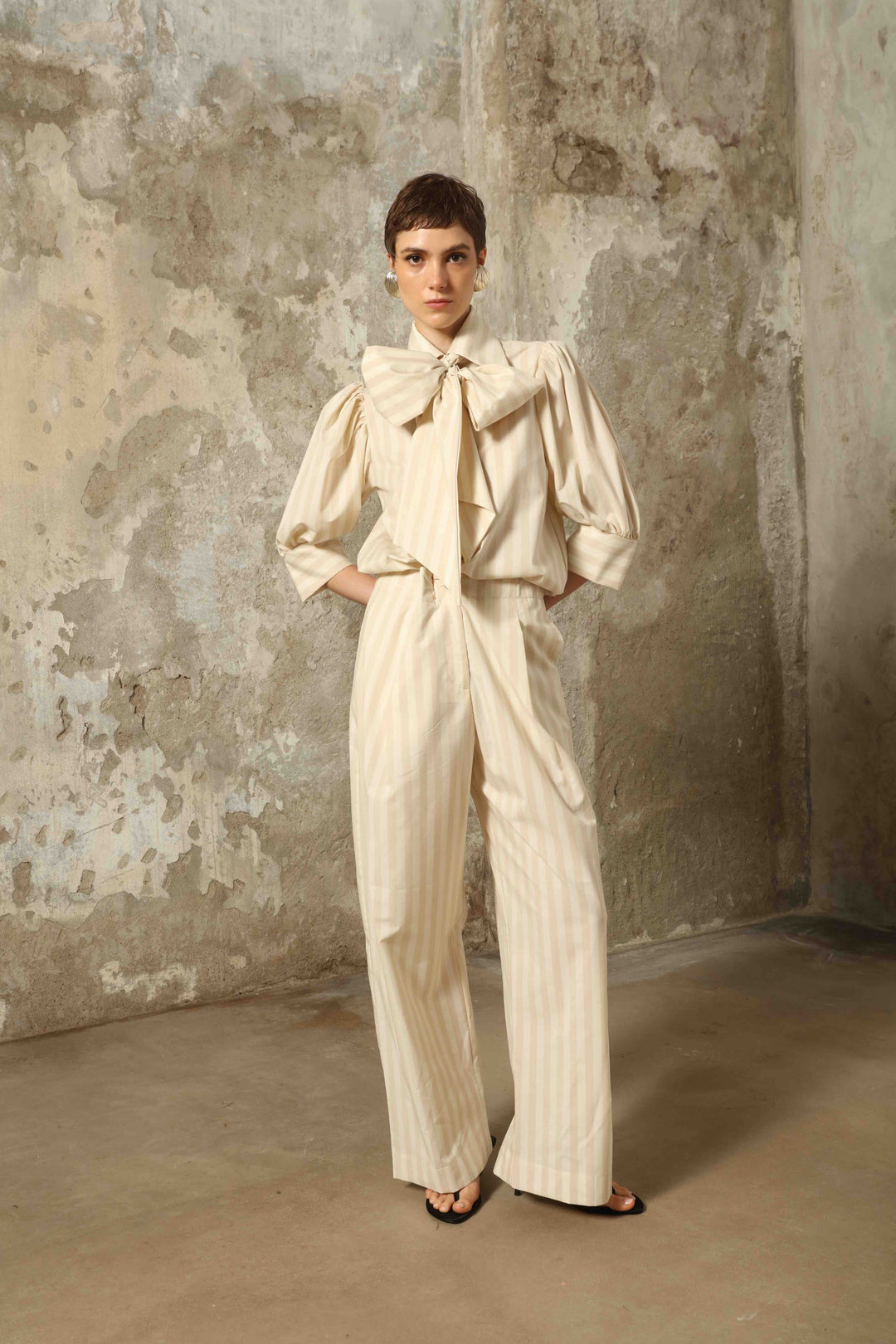 Model wearing the Gabriella Sand White Striped Trousers by House of Nomad Stories, featuring a high-rise waist, straight-leg tailored fit, and soft sand and white stripe pattern in a premium cotton, merino wool, and cashmere blend.