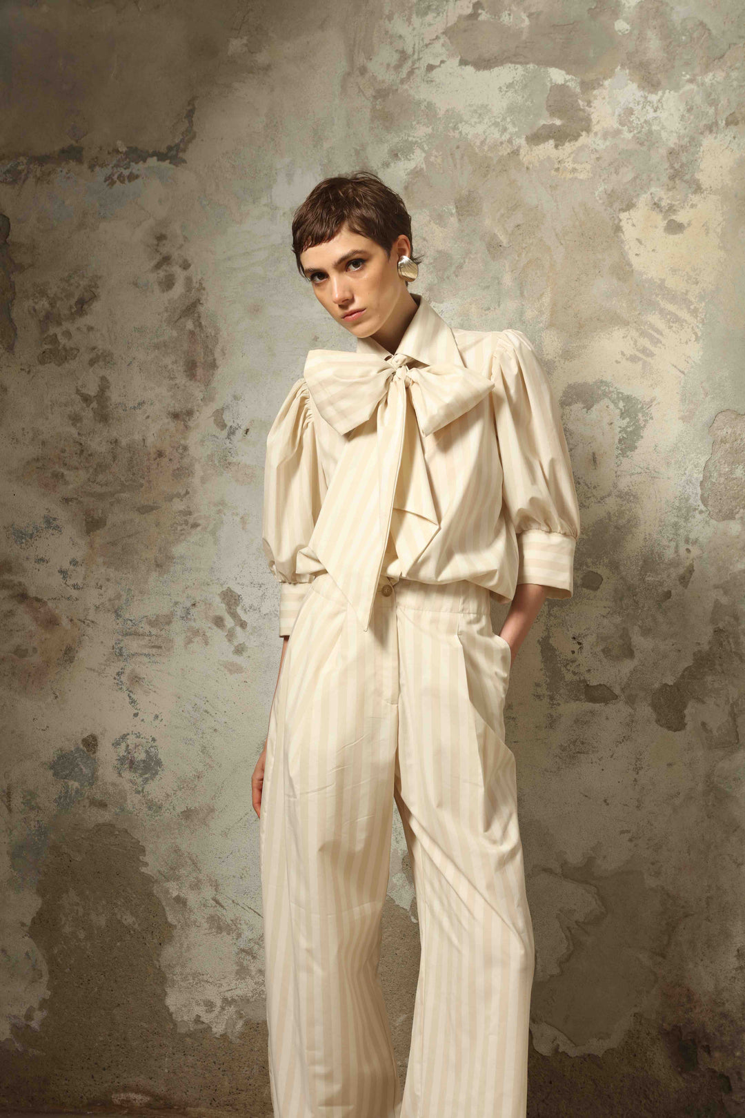 Model wearing the Gabriella Sand White Striped Trousers by House of Nomad Stories, featuring a high-rise waist, straight-leg tailored fit, and soft sand and white stripe pattern in a premium cotton, merino wool, and cashmere blend.