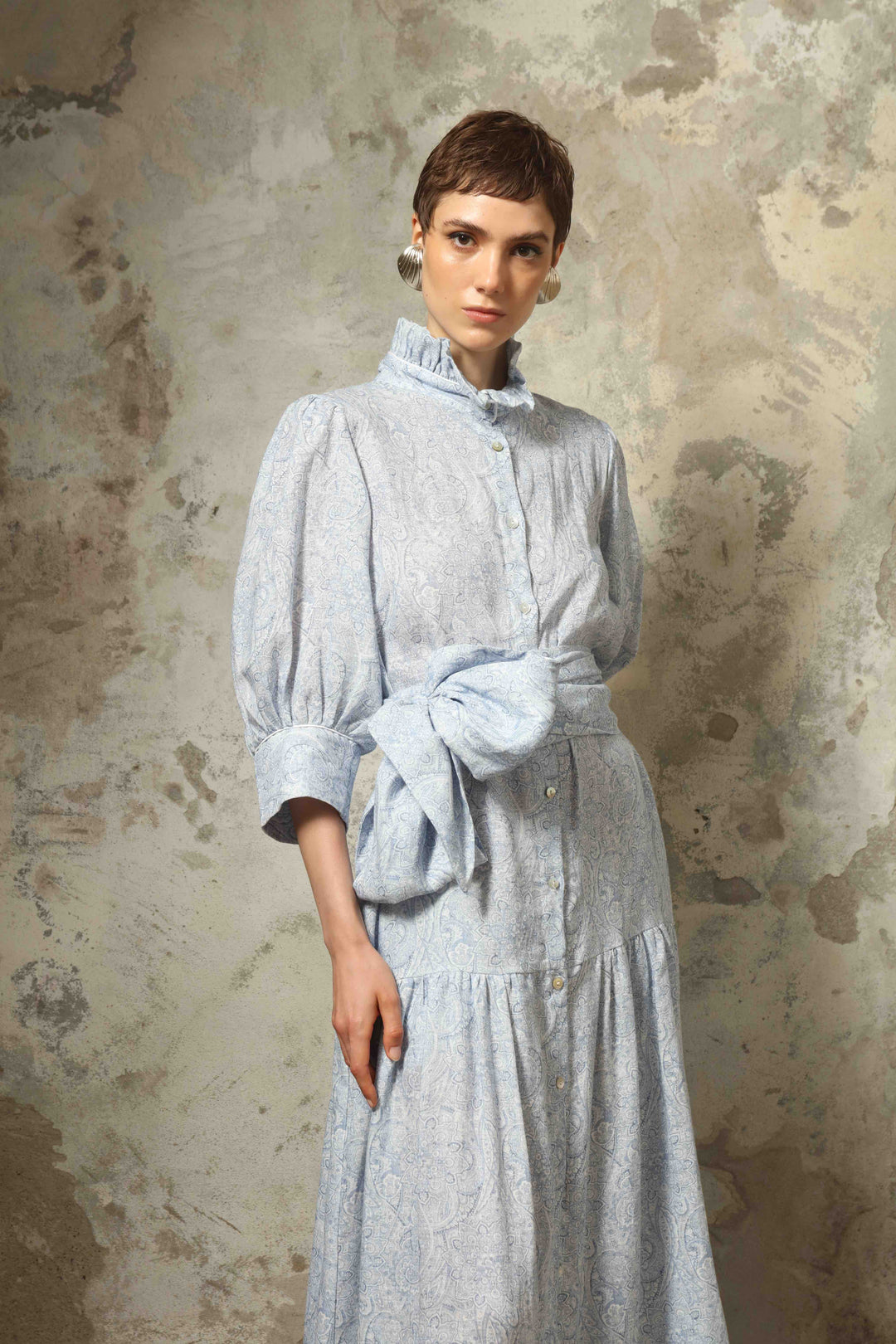 Model wearing the Karin Blue Paisley Dress by House of Nomad Stories, featuring 100% linen fabric, soft blue paisley print, and a relaxed flowing silhouette with breathable lightweight structure.