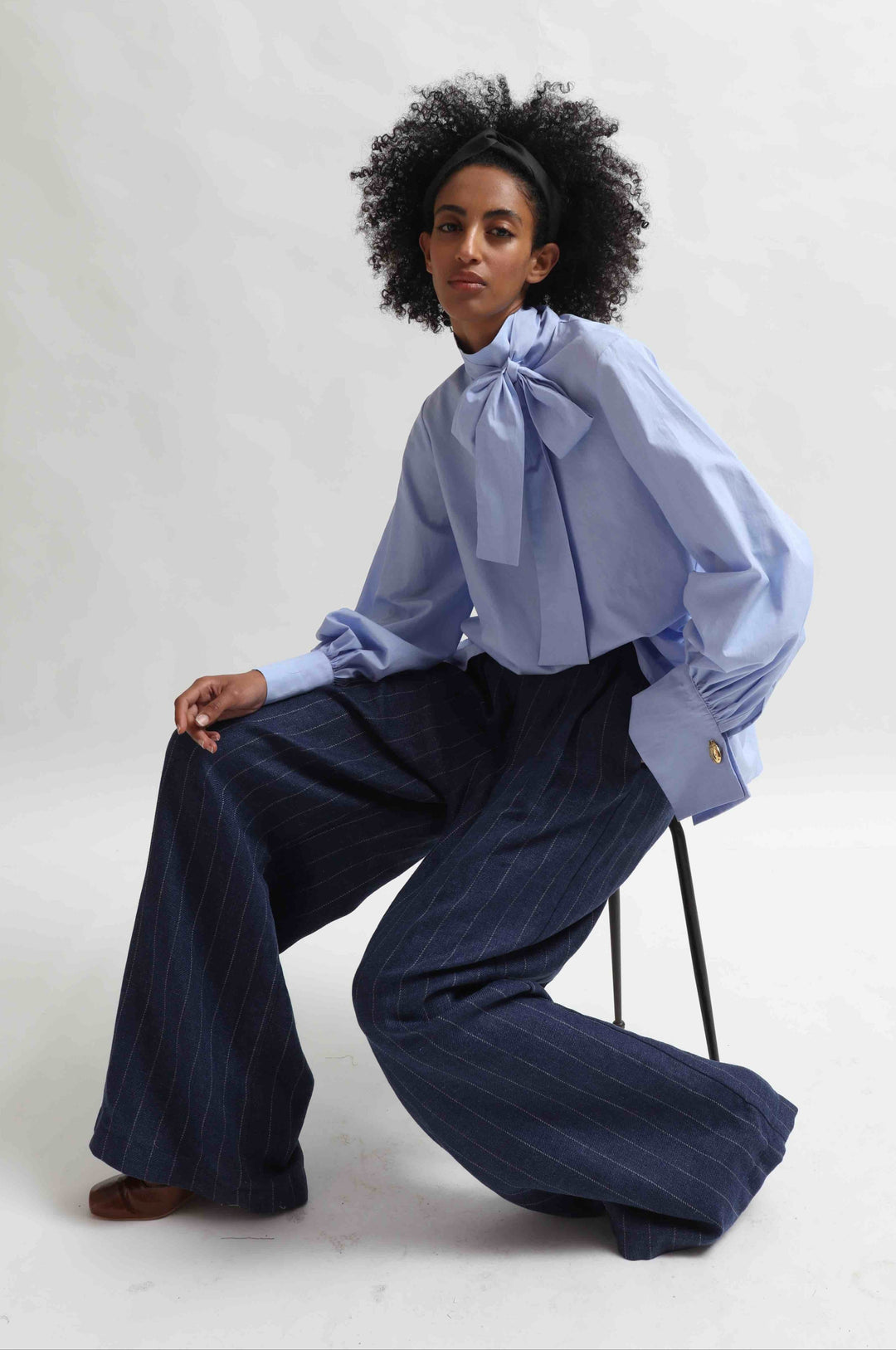 Model is wearing the Striped Navy Blue Parigi Trousers by House of Nomad Stories, crafted from a linen, wool, and silk blend with a relaxed wide-leg silhouette, subtle stripes, front pleats, and a mid-rise elasticated waistband.
