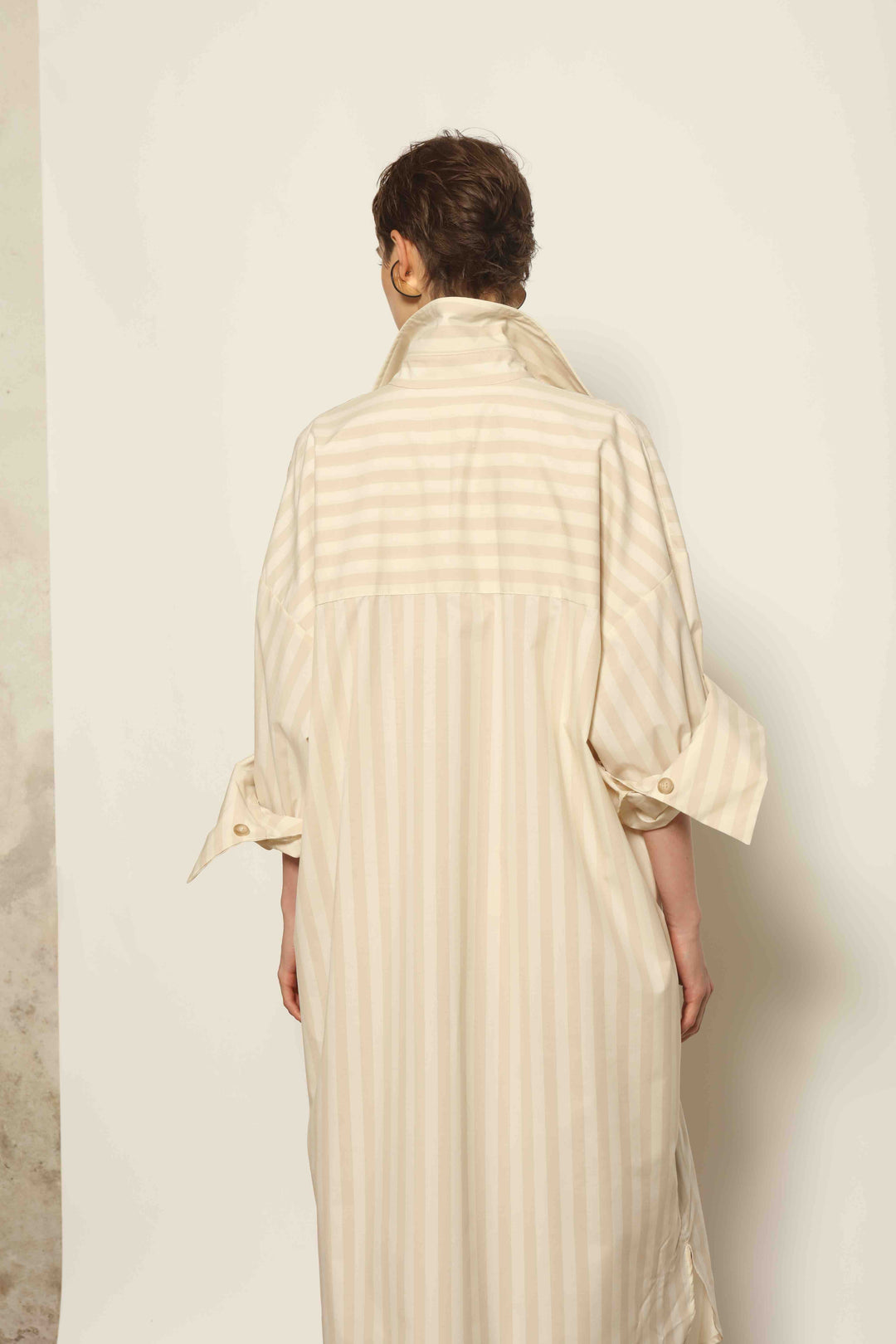 Model is wearing the Sand White Striped Bettina Dress by House of Nomad Stories, featuring a fluid silhouette with subtle sand and white vertical stripes and a soft, elegant drape.