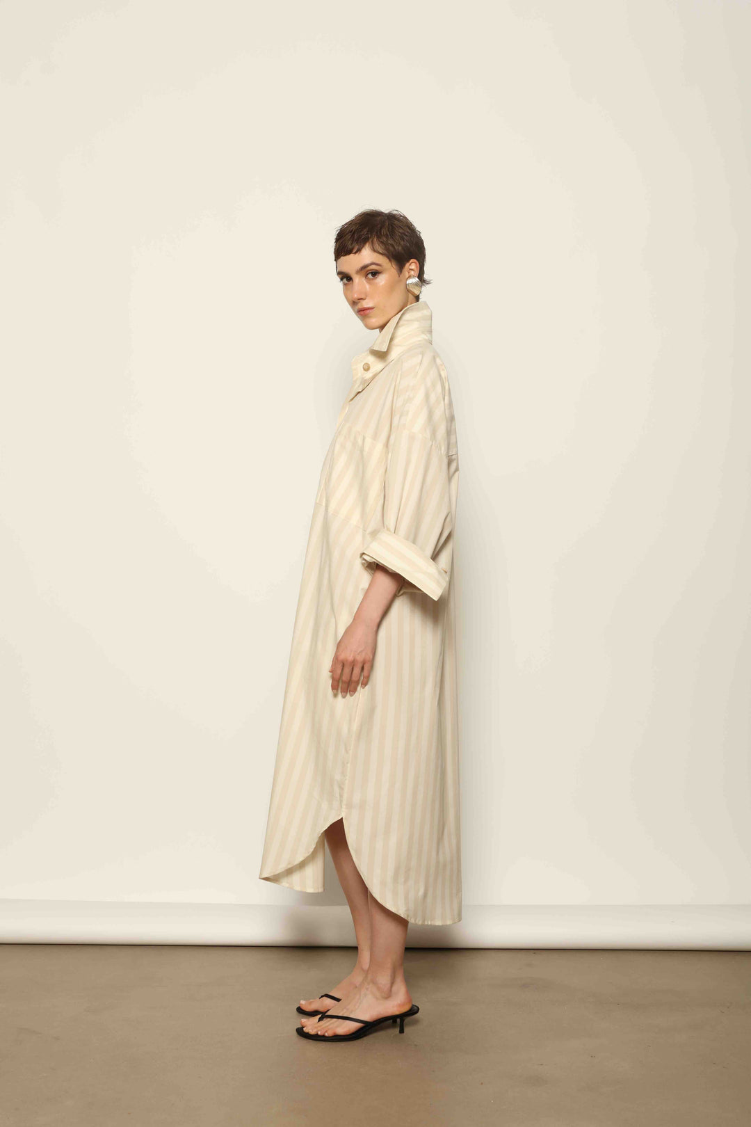 Model is wearing the Sand White Striped Bettina Dress by House of Nomad Stories, featuring a fluid silhouette with subtle sand and white vertical stripes and a soft, elegant drape.