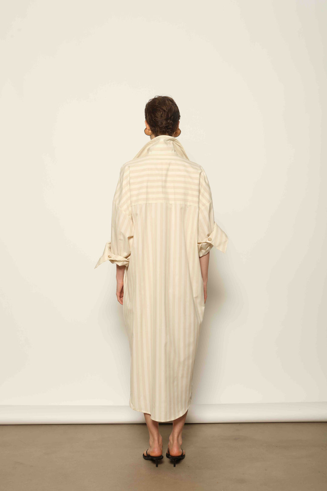 Model is wearing the Sand White Striped Bettina Dress by House of Nomad Stories, featuring a fluid silhouette with subtle sand and white vertical stripes and a soft, elegant drape.