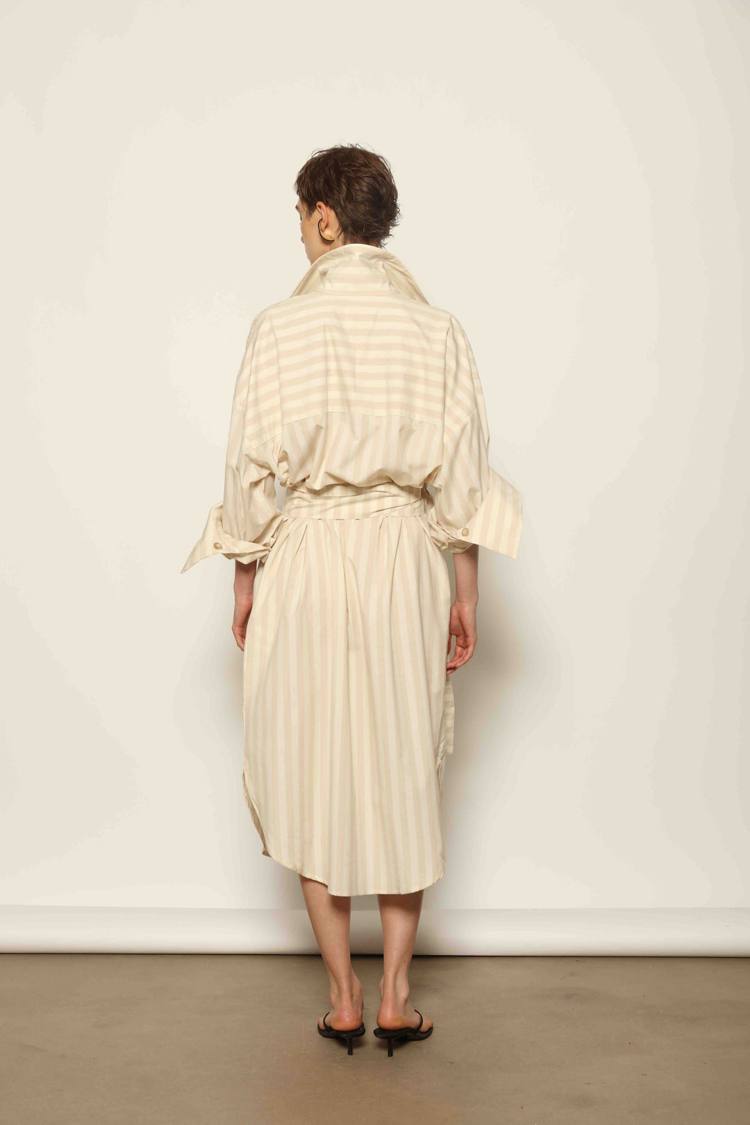 Model is wearing the Sand White Striped Bettina Dress by House of Nomad Stories, featuring a fluid silhouette with subtle sand and white vertical stripes and a soft, elegant drape.