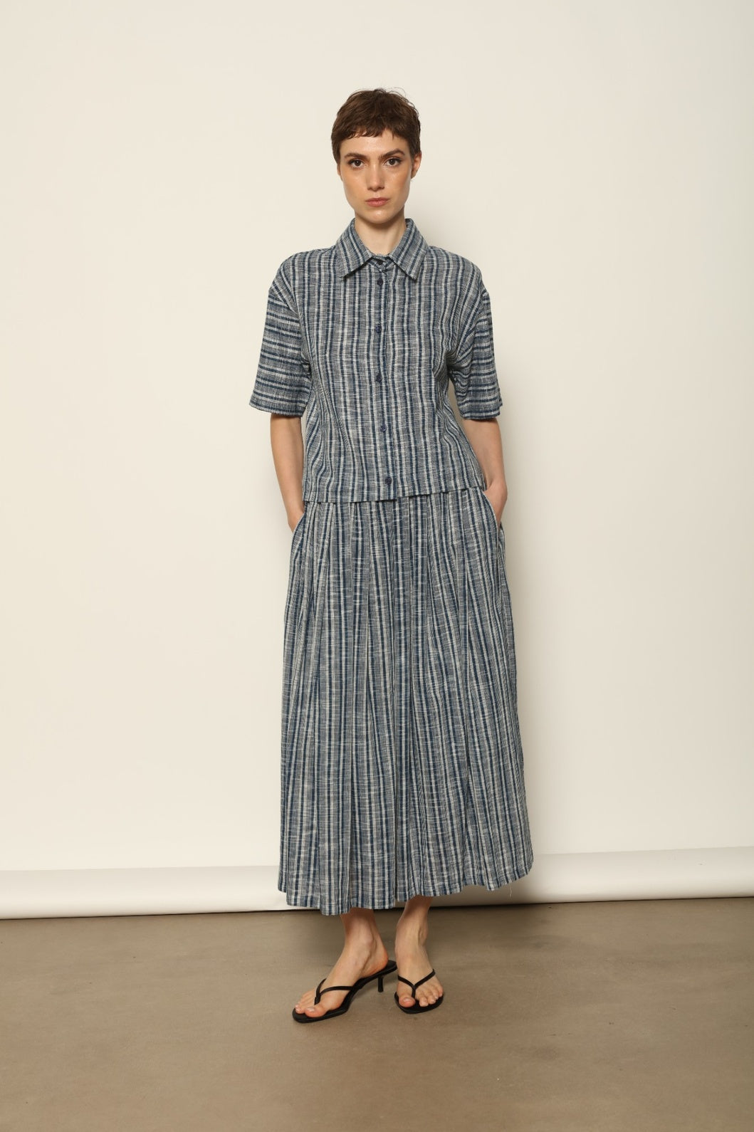 Model wearing the Seoul Indigo Blue Striped Ikat Midi Skirt by House of Nomad Stories, crafted from handwoven Kala cotton with an indigo blue and off-white ikat pattern, elasticated waistband, and relaxed midi silhouette.