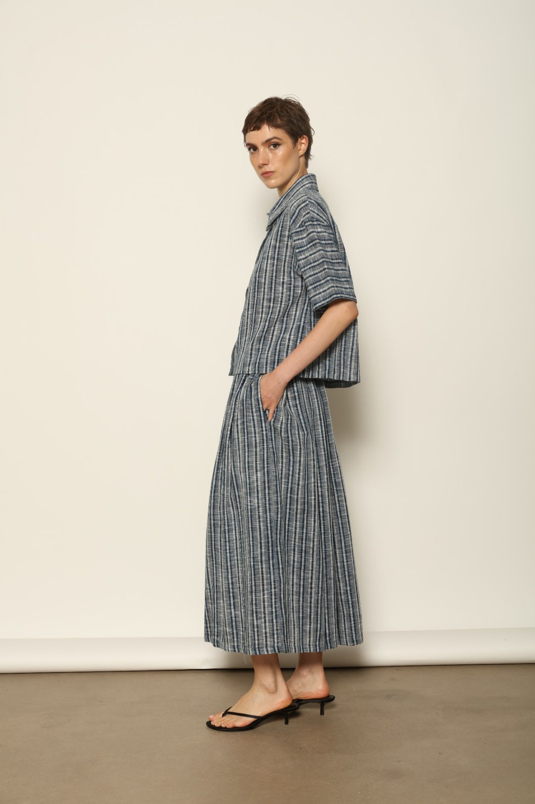Model wearing the Seoul Indigo Blue Striped Ikat Midi Skirt by House of Nomad Stories, crafted from handwoven Kala cotton with an indigo blue and off-white ikat pattern, elasticated waistband, and relaxed midi silhouette.