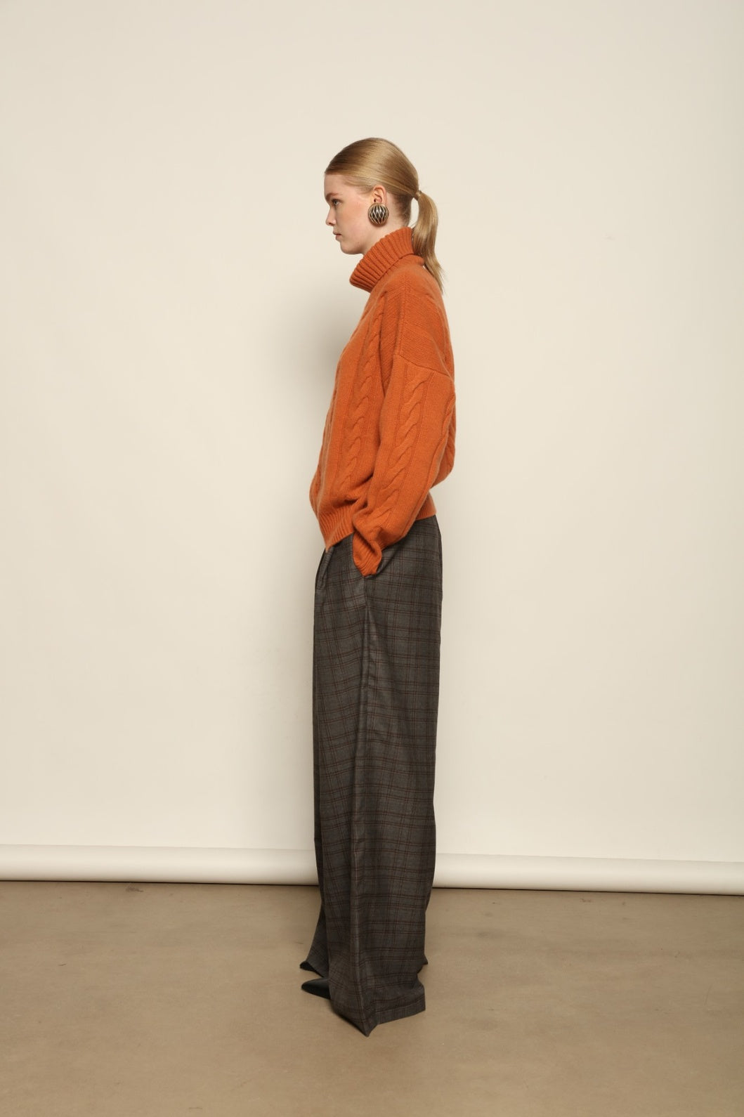 Model is wearing the Brick Noorah Turtleneck Sweater by House of Woven Stories, a pure ethically sourced cashmere cable-knit sweater in a rich brick tone with ribbed high collar and cuffs.