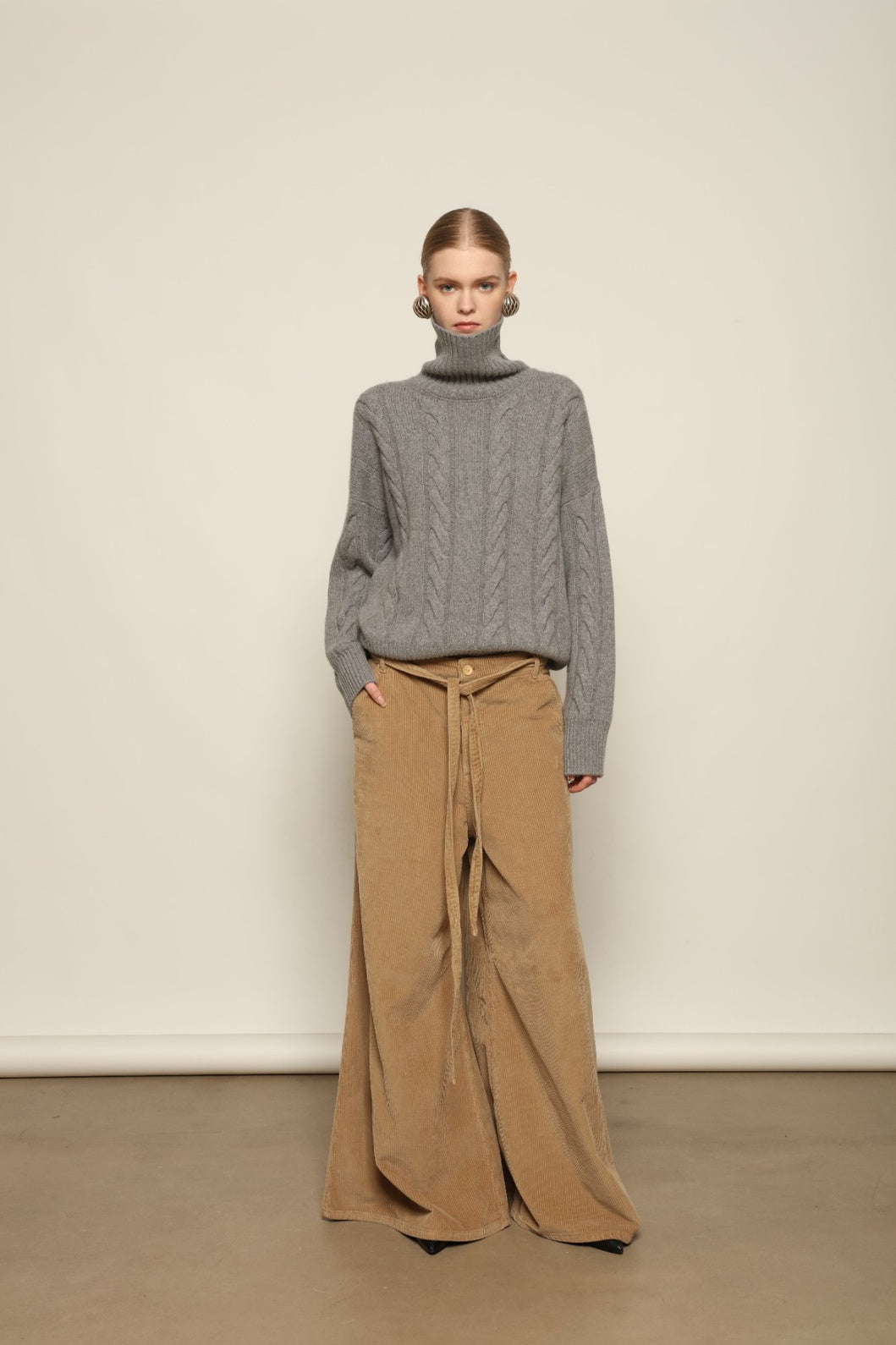 Model is wearing the Graphite Noorah Turtleneck Sweater by House of Woven Stories, a pure ethically sourced cashmere cable-knit sweater with wide ribbed turtleneck and cuffs in graphite.