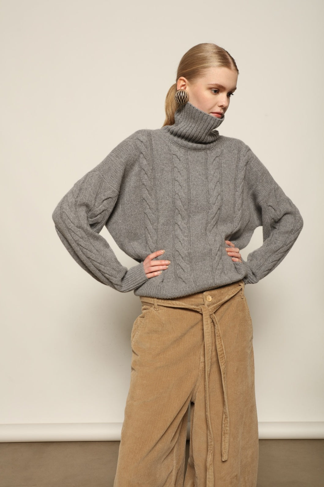 Model is wearing the Graphite Noorah Turtleneck Sweater by House of Woven Stories, a pure ethically sourced cashmere cable-knit sweater with wide ribbed turtleneck and cuffs in graphite.