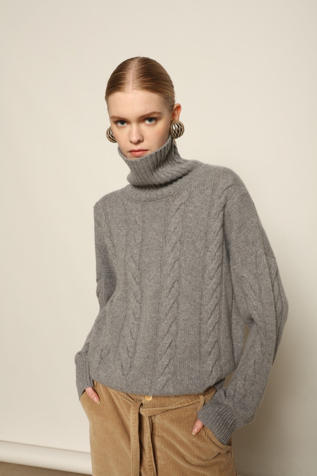 Model is wearing the Graphite Noorah Turtleneck Sweater by House of Woven Stories, a pure ethically sourced cashmere cable-knit sweater with wide ribbed turtleneck and cuffs in graphite.