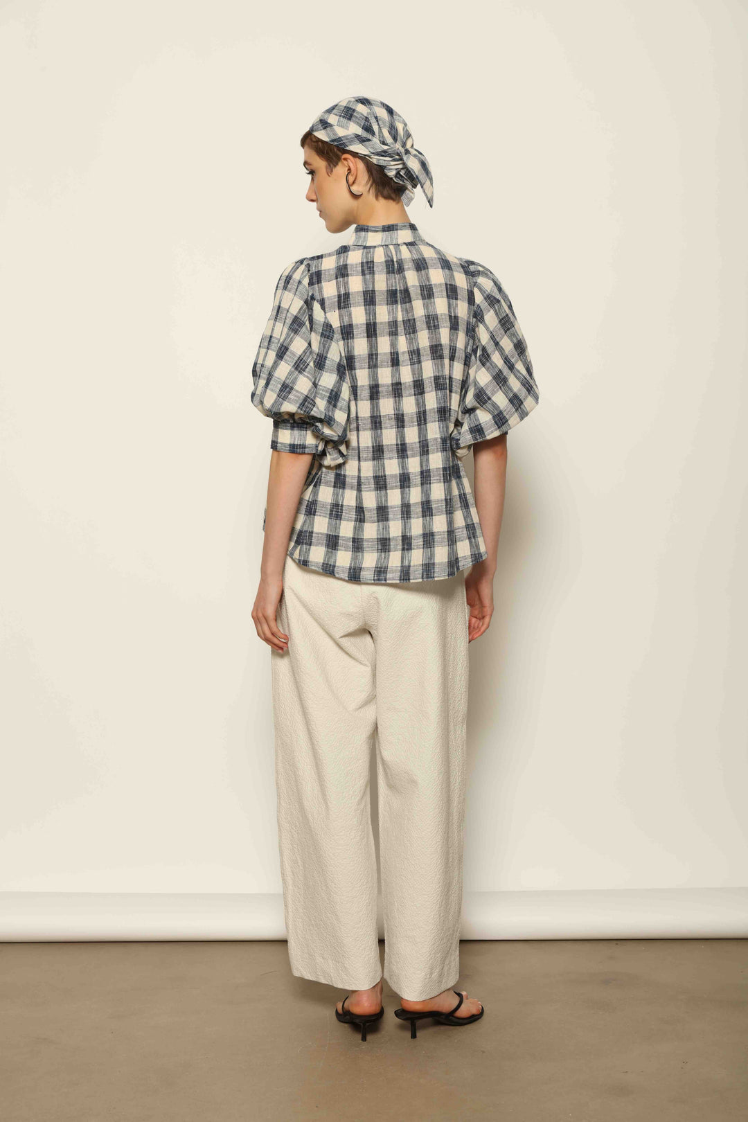 Model wearing the Lola Check Blouse by House of Woven Stories, crafted from 100% handloomed Kala cotton with a blue and white check pattern, Mandarin collar, button-front closure, and balloon sleeves.
