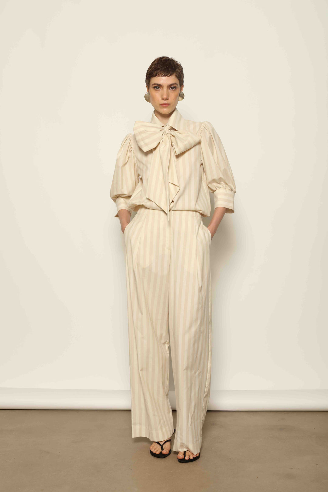 Model wearing the Karin Sand White Macro-Striped Blouse by House of Nomad Stories, featuring a relaxed fit, sand and white macro stripes, classic collar, and soft blend of cotton, merino wool, and cashmere.