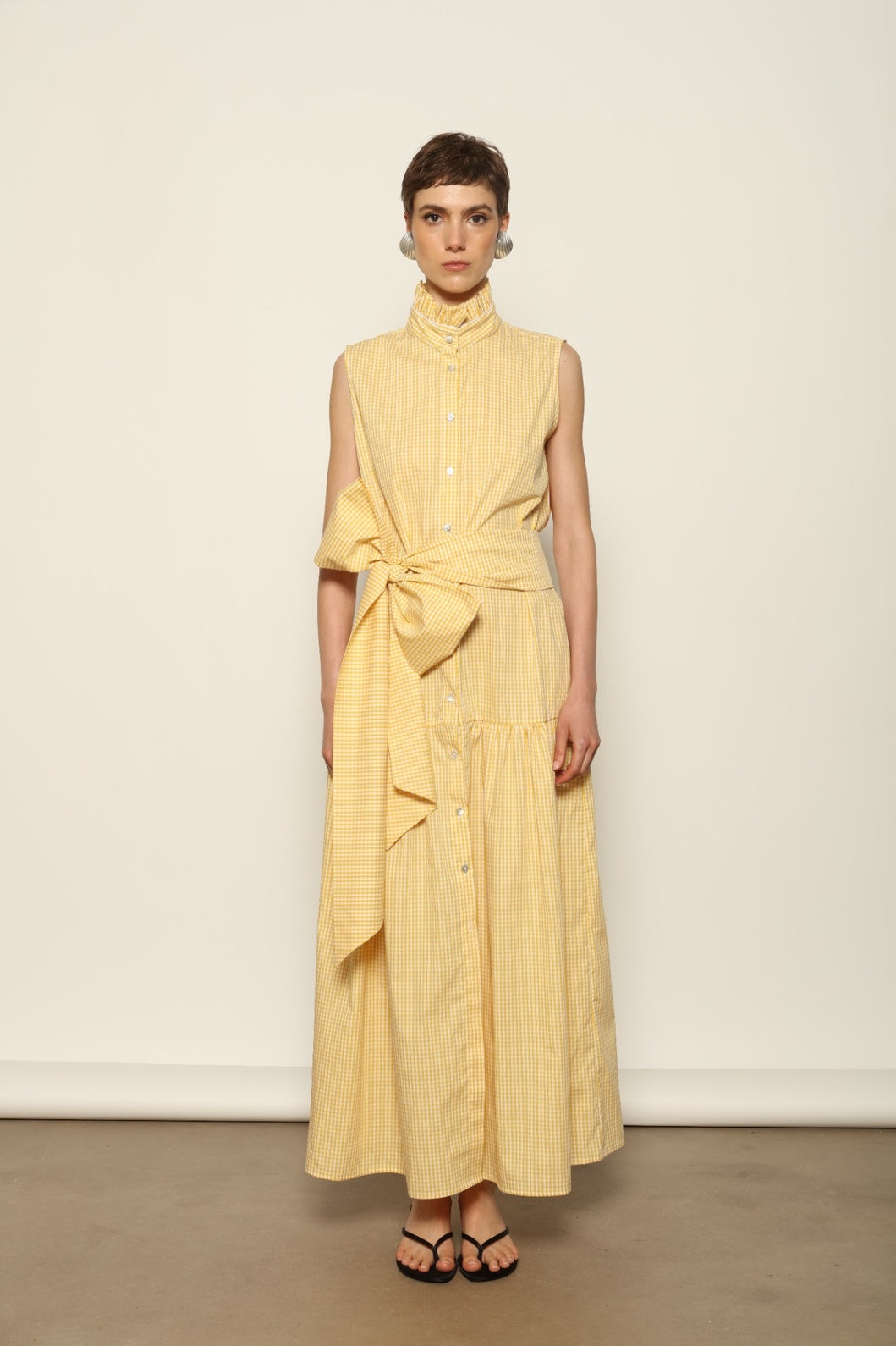 Model wearing the Karin Vichy Butter Yellow Dress by House of Woven Stories, featuring 100% organic cotton fabric, butter yellow gingham pattern, sleeveless silhouette, signature ruffled collar, and white piping details.