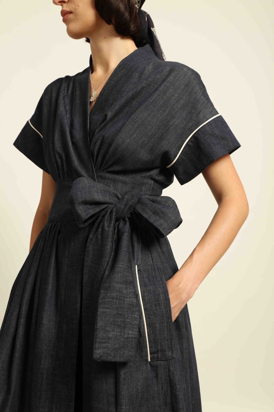 Model is wearing the Saint Remy wrap dress by House of Woven Stories in 100% cotton, featuring a denim-blue color with white pinstripes, drop shoulders, short sleeves, deep V-neckline, and self-tie belt, handmade in Italy.