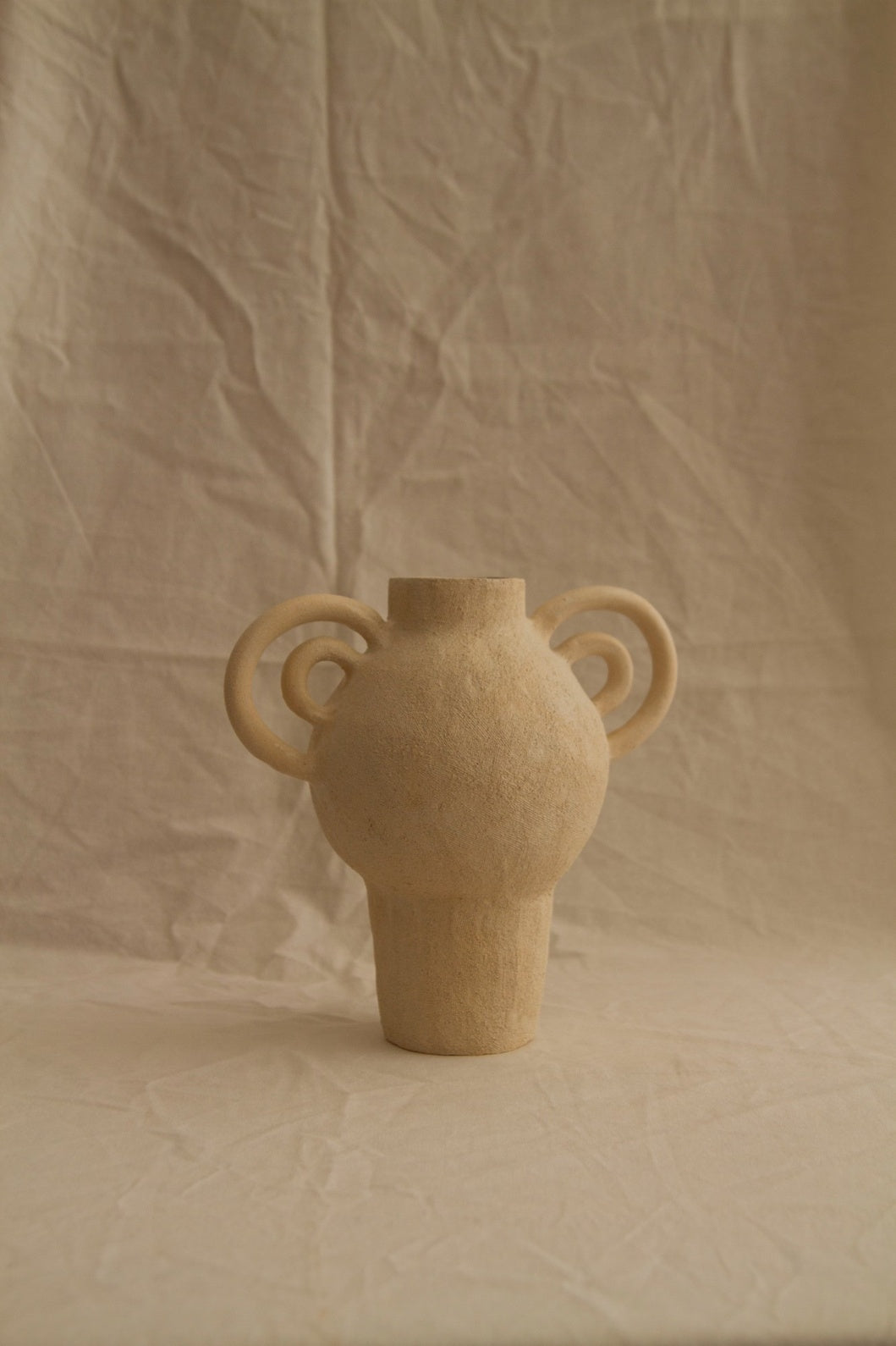 Beige ceramic vase with two handles on a beige textured background. L’Enfant Lune aux Doubles Anses by Clandestine Céramique, handbuilt in sandy clay with double curving handles and soft glaze, part of the House of Nomad Stories selection.