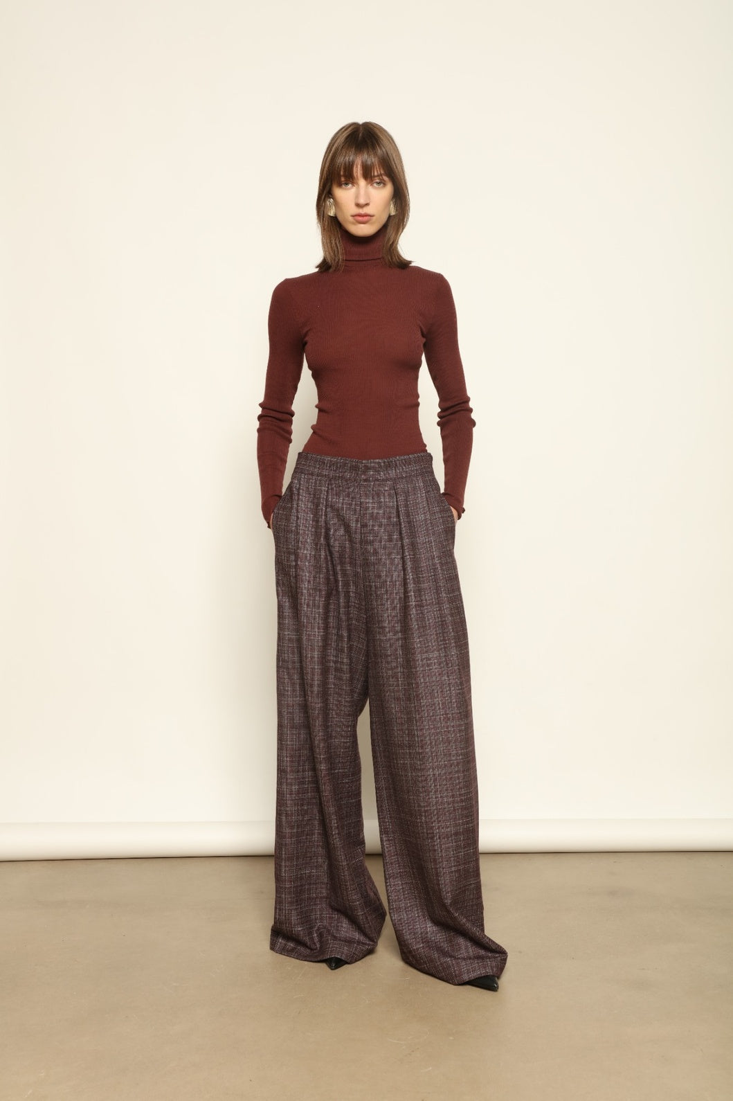 Model is wearing the Parigi Burgundy Lana Trousers by House of Woven Stories – Italian-tailored trousers crafted from a luxurious blend of merino wool, silk, and linen, featuring front pleats, side pockets, and a relaxed yet elegant burgundy silhouette.
