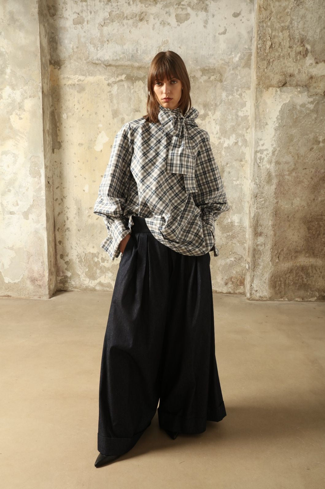 Model is wearing the Regula Maxi Check Popeline Cotton Blouse by House of Nomad Stories, crafted from crisp poplin cotton in an oversized check pattern with a waist tie and buttoned cuffs.