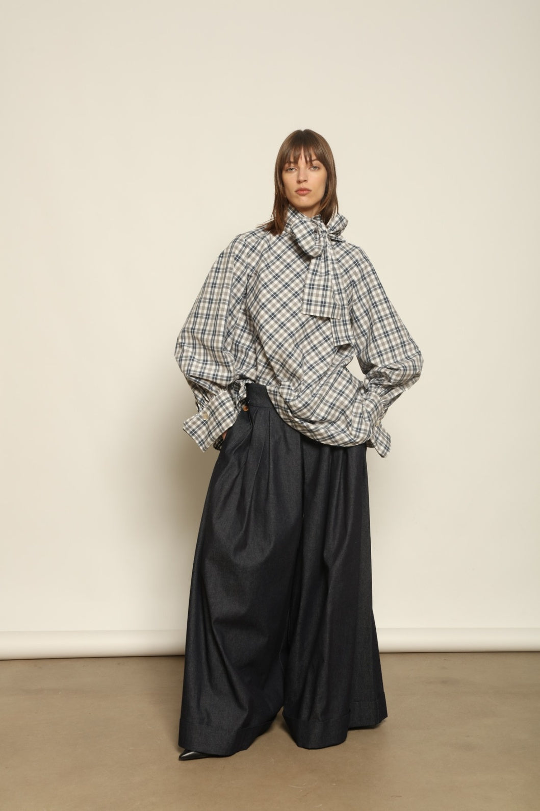 Model is wearing the Regula Maxi Check Popeline Cotton Blouse by House of Nomad Stories, crafted from crisp poplin cotton in an oversized check pattern with a waist tie and buttoned cuffs.