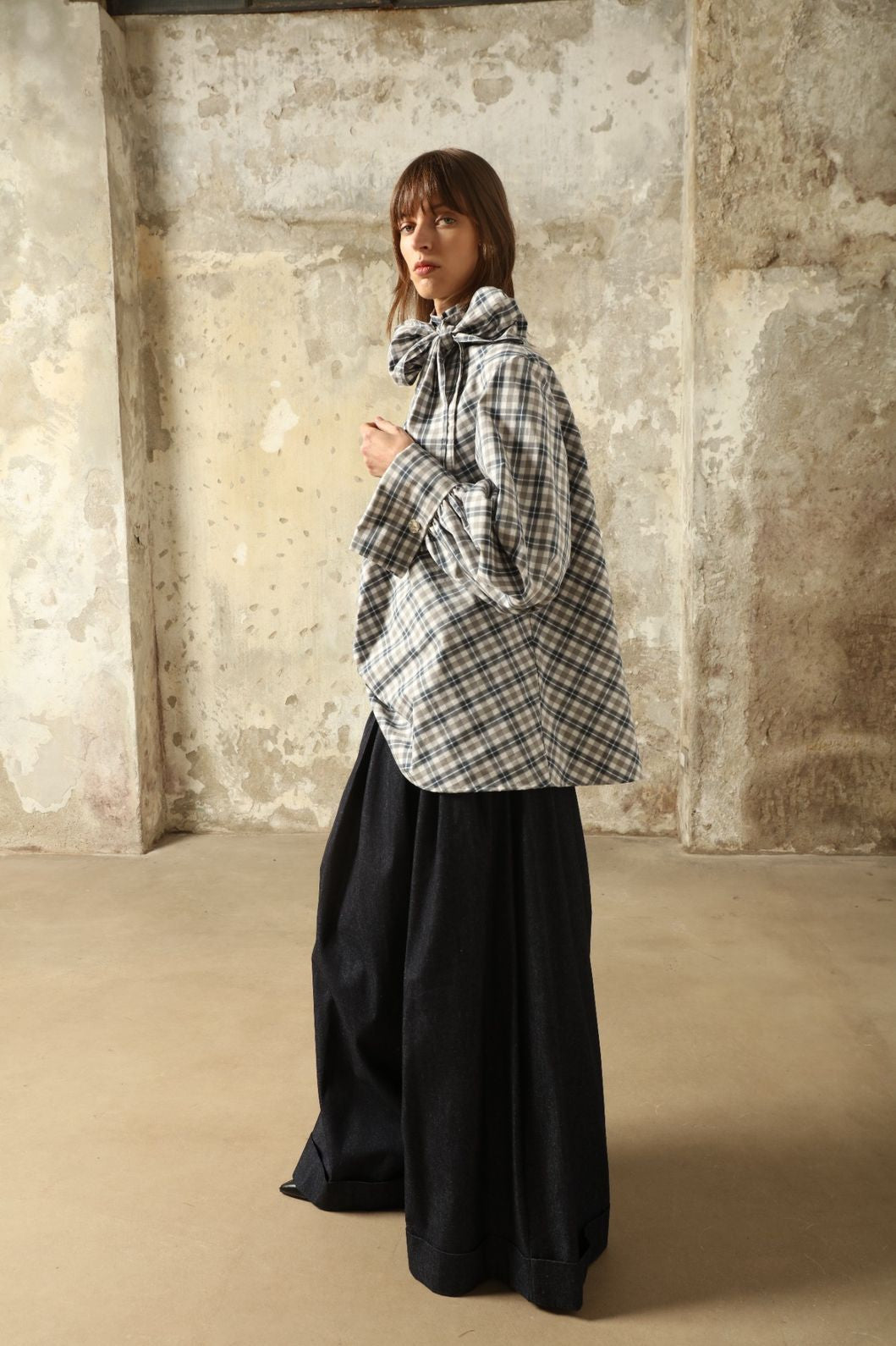Model is wearing the Regula Maxi Check Popeline Cotton Blouse by House of Nomad Stories, crafted from crisp poplin cotton in an oversized check pattern with a waist tie and buttoned cuffs.